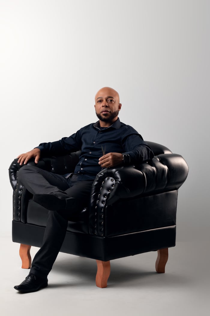 Confident bald man in a studio setting sitting on a black leather armchair wearing formal attire.